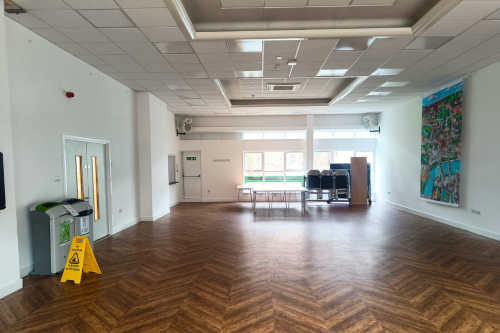 The main hall is a large rectangular space with wooden flooring and windows at one end. There is a piece of art hanging on the wall on the left wall and double doors on the right wall. 